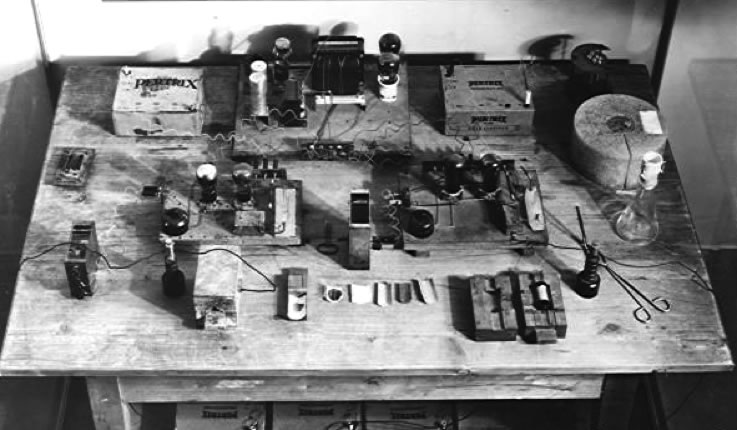 La table de travail d’Otto Hahn en 1938 ©Hulton Archives. Au premier plan, de gauche à droite, un compteur numérique, un amplificateur et un compteur Geiger, au second plan une batterie (d’autres sous la table) et à droite une source de neutrons entouré de paraffine.